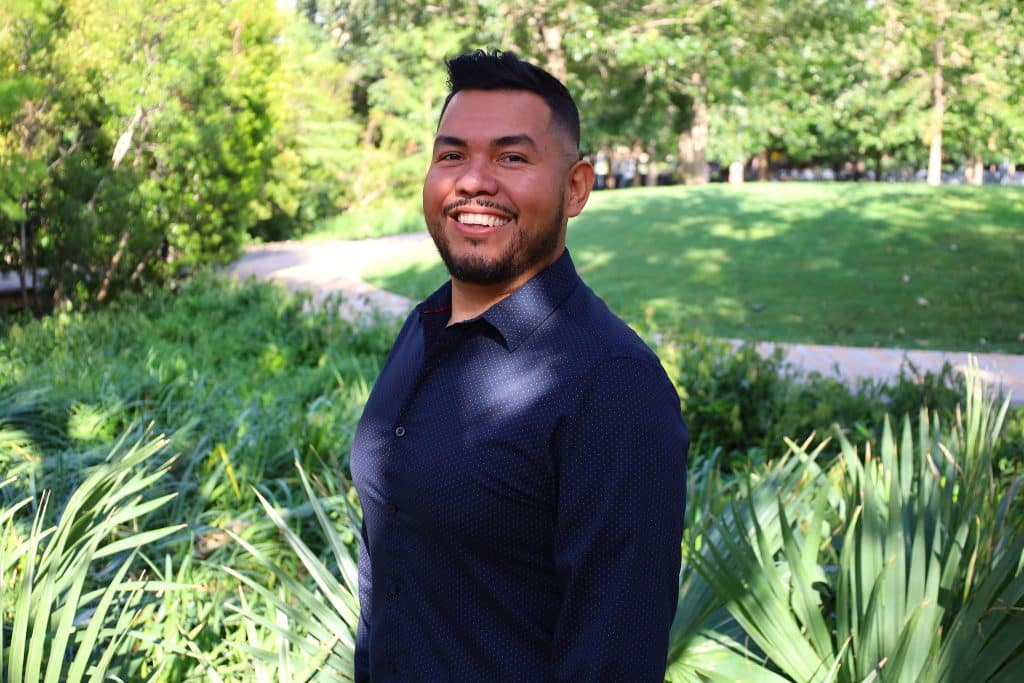 Hugo Rivera, smiling in a navy shirt, standing amidst greenery in a park, representing BridgeYear's commitment to supporting students from underserved communities.