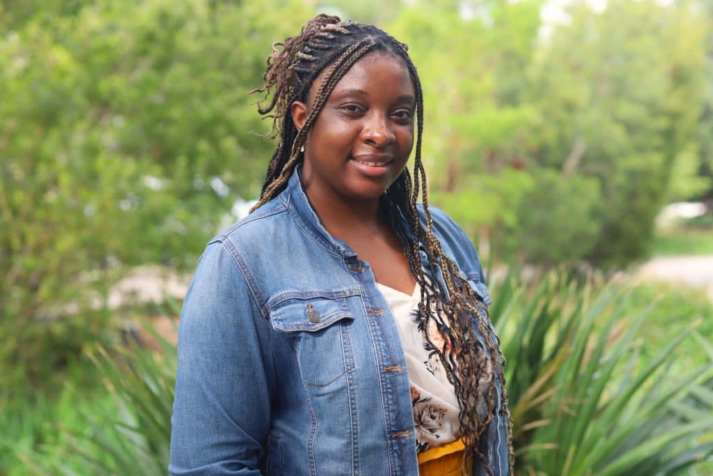 Portrait of a woman with braided hair wearing a denim jacket, smiling in front of greenery, representing the dedicated team at BridgeYear supporting students from underserved communities.