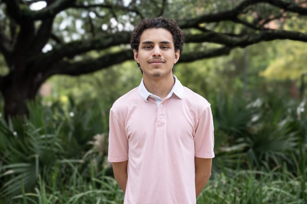 Young man in a pink polo shirt standing outdoors among greenery, representing the BridgeYear team dedicated to supporting students from underserved communities.