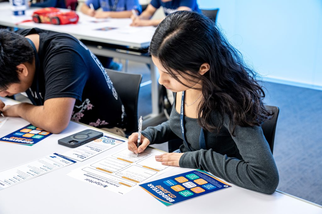 Students participating in the BridgeYear ES3 Conference, focusing on resume building and professional skills, with one girl writing on a worksheet and a boy in the background engaging in the session.